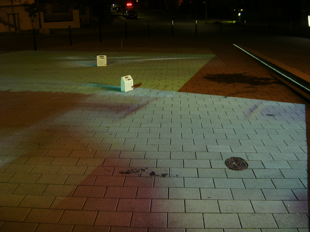 Zwei kleine beleuchtete Kisten auf einem nächtlichen Platz neben Straßenbahnschienen.