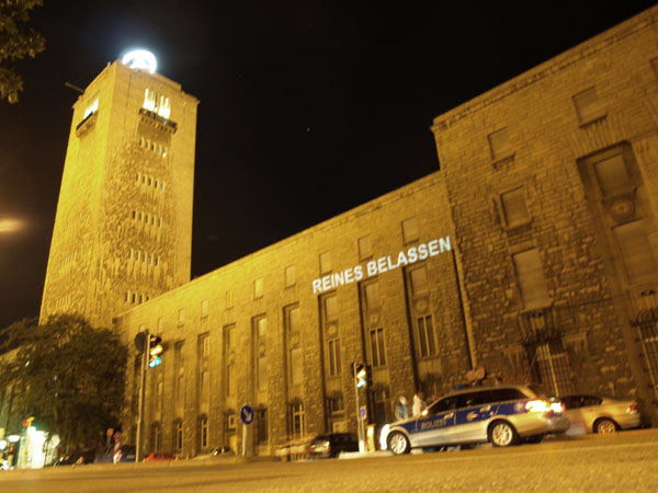 Die Worte „REINES BELASSEN“ werden nachts auf eine Gebäudefassade mit Turm projiziert.