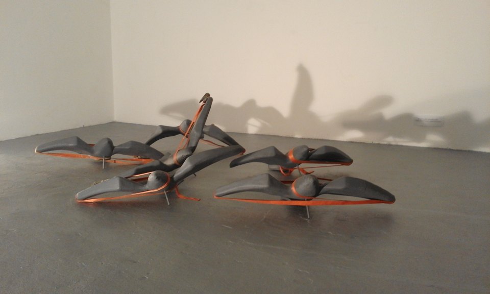 Seitliche Ansicht der umgekippten grauen Sitzformen mit orangefarbenen Gurten; die Formen werfen Schatten an die Wand.
