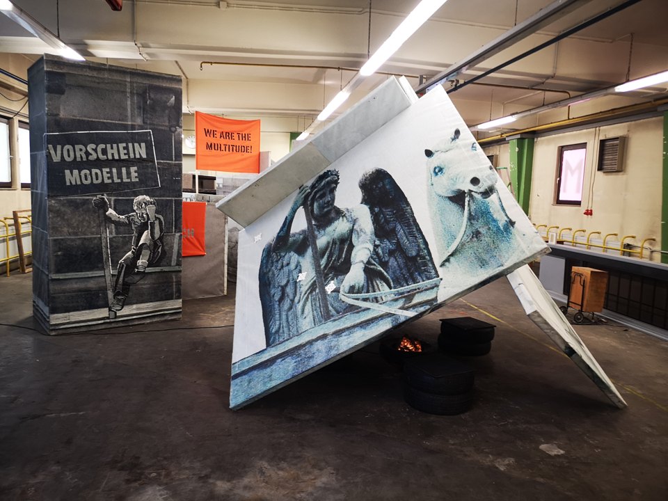 Schräg aufgestellte Figurentafel neben bedruckten Architekturflächen; eine Leiter und Textplakate stehen dahinter.