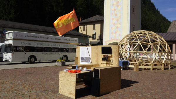 Holzstand mit orangefarbener Fahne auf einem Platz; dahinter stehen ein Bus und eine kuppelförmige Konstruktion.