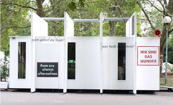 Weißer Pavillon mit hohen Wandflächen, Durchgängen und Schriftplakaten unter Bäumen.