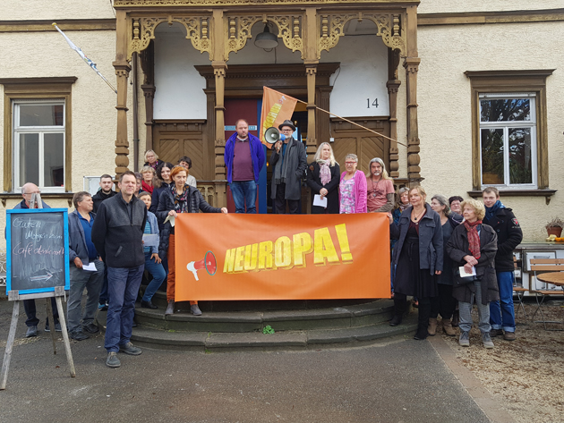 Gruppenfoto vor einem Gebäude; die Gruppe hält ein orangefarbenes Banner „NEUROPA!“.