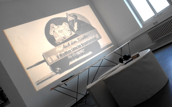 Historische Abbildung mit Schrift wird auf eine Wand projiziert; davor stehen Möbel und technische Geräte.