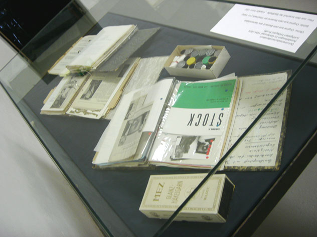 Glasvitrine mit Heften, handschriftlichen Dokumenten, einer kleinen Schachtel und einer Texttafel.