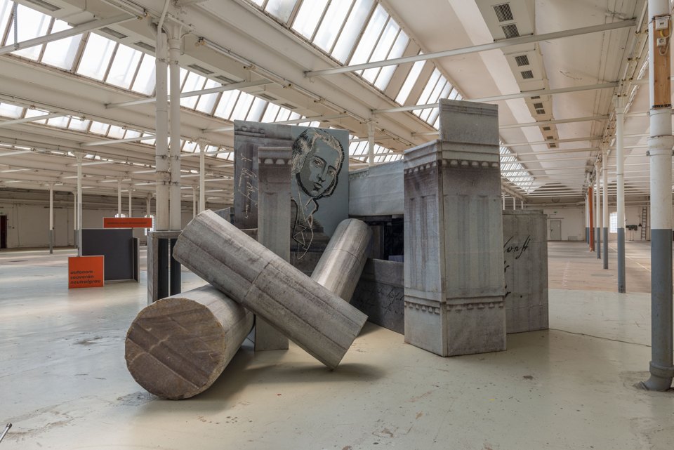 Installation aus bedruckten Architekturflächen und zwei schräg liegenden Säulenformen in einer Halle.