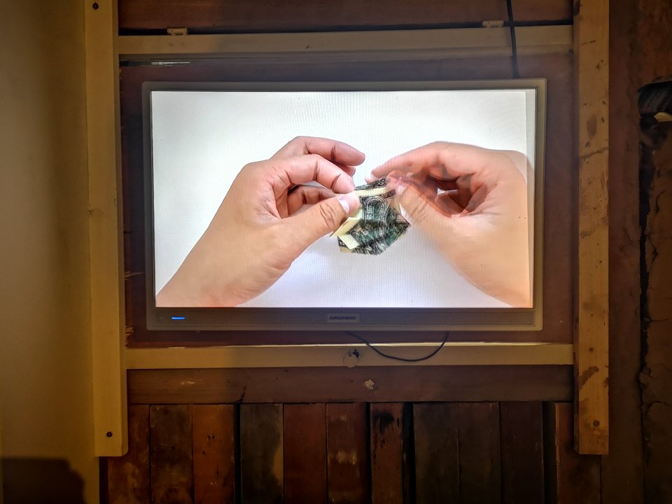 Monitor in einer Holzkonstruktion zeigt zwei Hände beim Bearbeiten eines kleinen Gegenstands.