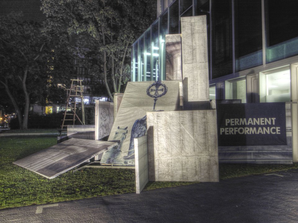 Seitliche nächtliche Ansicht der Architekturflächen mit einer Figurendarstellung und dem Schild „PERMANENT PERFORMANCE“.
