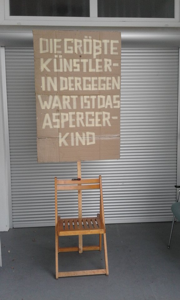 Pappschild über einem Holzstuhl mit der Aufschrift „DIE GRÖßTE KÜNSTLER-IN DER GEGEN-WART IST DAS ASPERGER-KIND“.