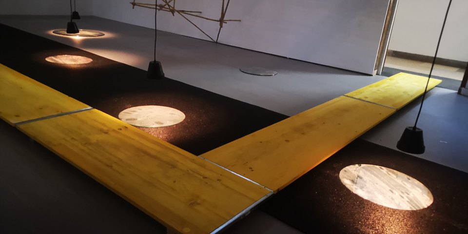 Gelber Laufsteg durch einen dunklen Ausstellungsraum mit beleuchteten kreisförmigen Szenen.