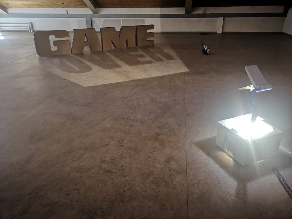 Installation aus großen Buchstaben mit Bodenschrift „OVER“; rechts steht eine kleine beleuchtete Konstruktion.