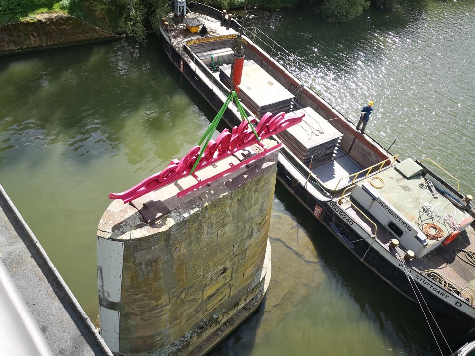 Blick von einer Brücke auf den Pfeiler mit rotem Schriftzug und ein daneben fahrendes Schiff.