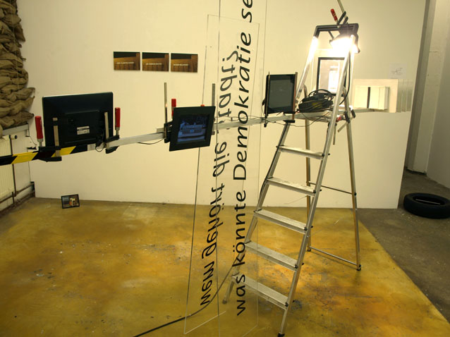 Installation mit Leiter, Bildschirmen, einem hohen beschrifteten Element und seitlich befestigten Gegenständen.