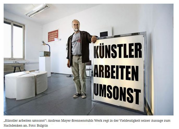 Person steht neben einem großen Leuchtkasten mit der Aufschrift „KÜNSTLER ARBEITEN UMSONST“.