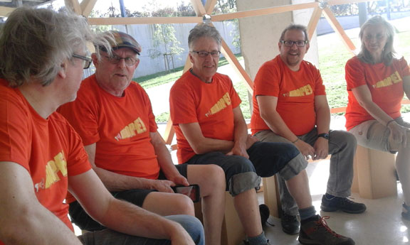 Fünf Personen in orangefarbenen T-Shirts sitzen nebeneinander in einem hellen Innenraum.