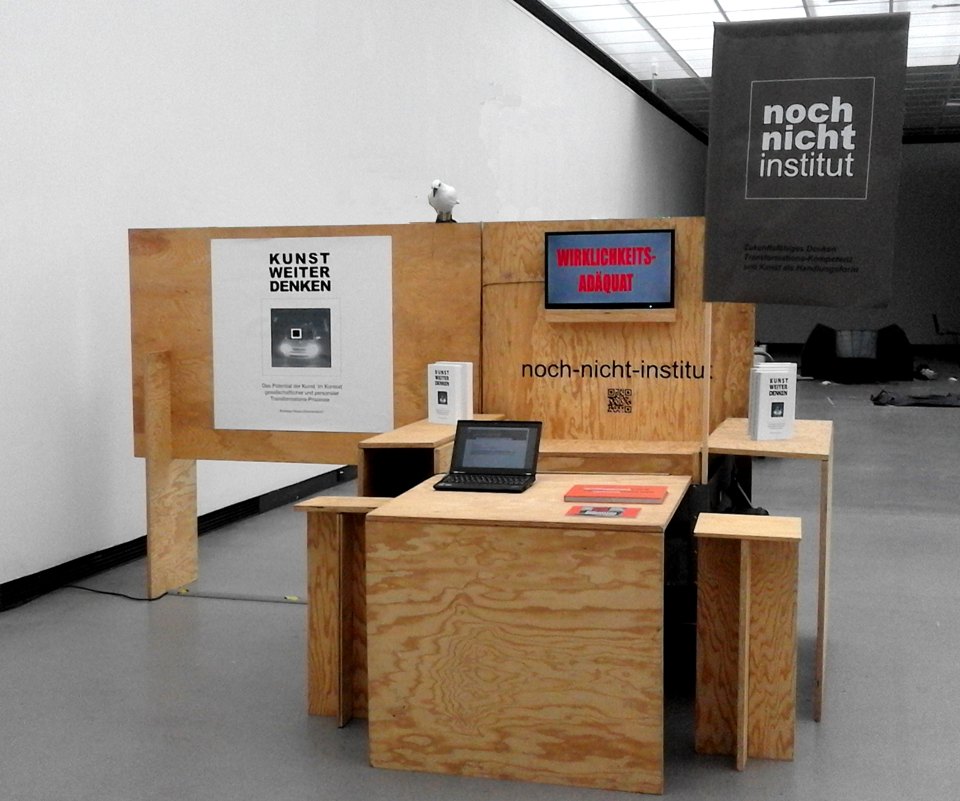 Frontansicht eines Informationsstands aus Holz mit Tisch, Büchern, Bildschirm und Schildern des noch-nicht-instituts.