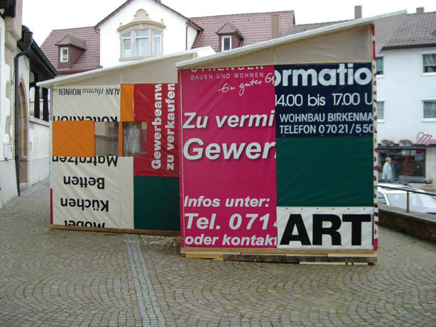 Seitliche Ansicht der hausartigen Konstruktion aus Texttafeln und einer offenen Tür auf einem Platz.