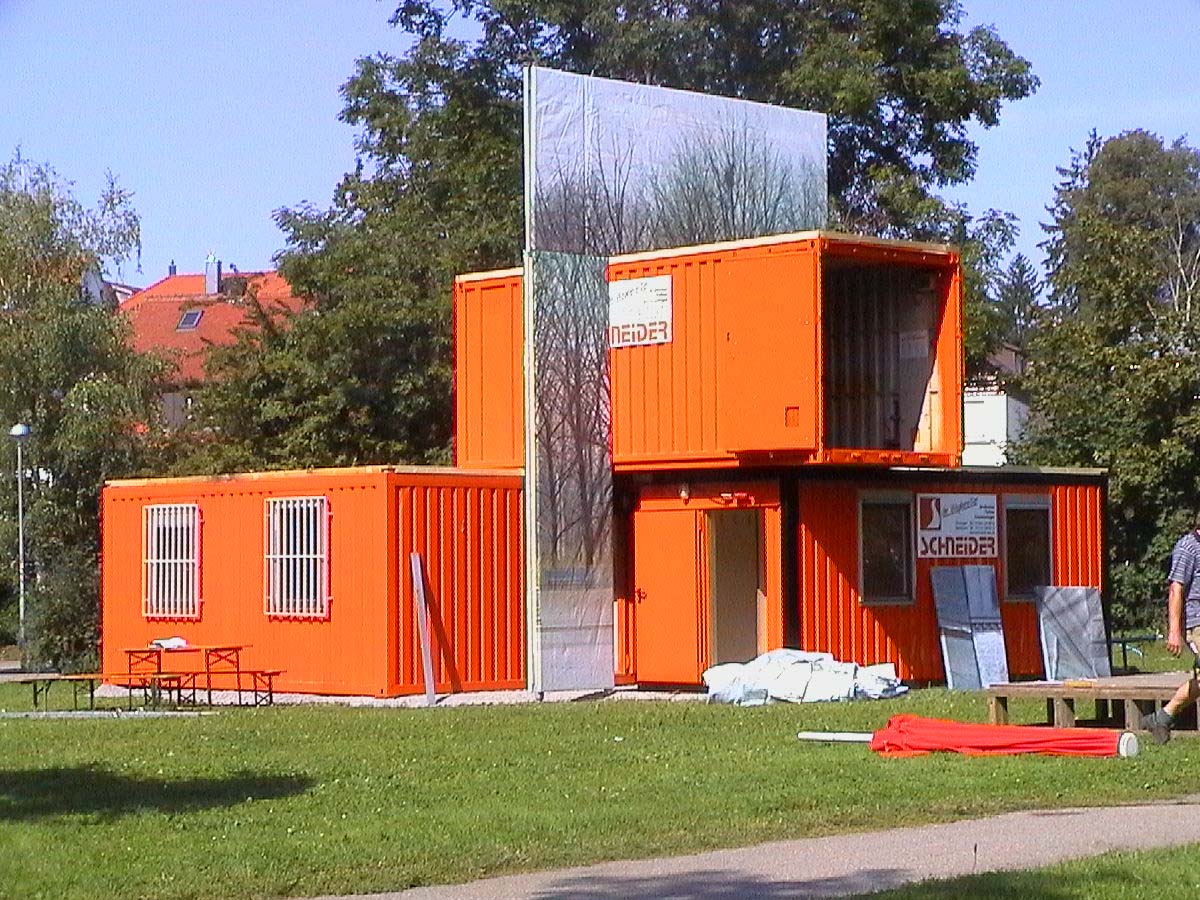 Gestapelte orangefarbene Container mit einem hohen verkleideten Wandelement auf einer Wiese.