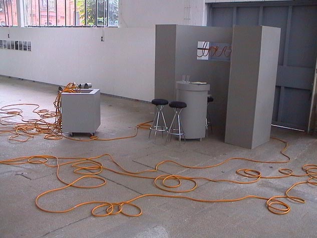 Orangefarbene Seile verlaufen in Schlaufen über den Boden eines Ausstellungsraums mit Tisch, Hockern und Wandaufbau.