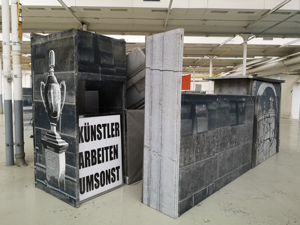 Bedruckte Architekturflächen mit einer Vasenabbildung und dem Schild „KÜNSTLER ARBEITEN UMSONST“.
