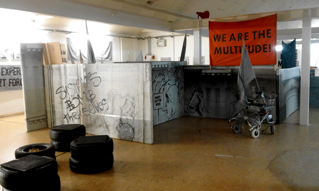 Bedruckte Architekturflächen mit Schrift „SYSTEMS BOMB themselves“, Reifen und Rollator unter einem orangefarbenen Banner.