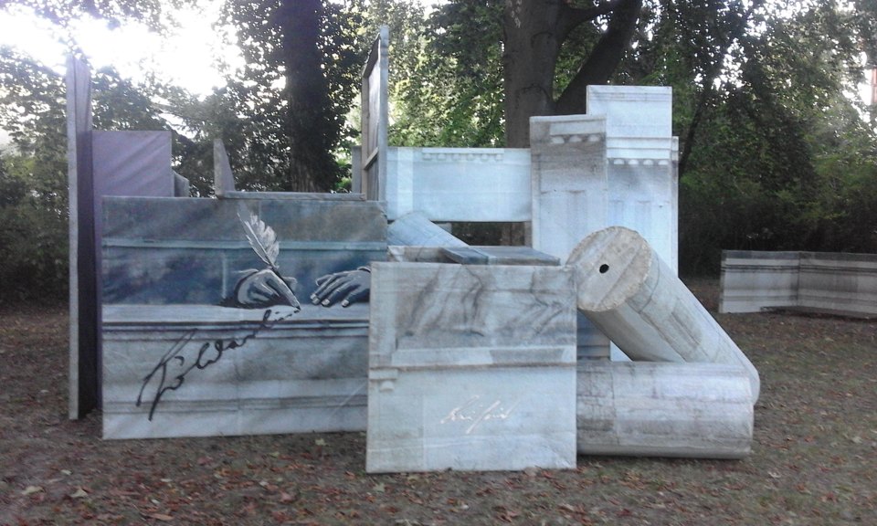 Seitliche Ansicht der Installation aus bedruckten Quadern, Tafeln und liegenden Säulenformen im Freien.