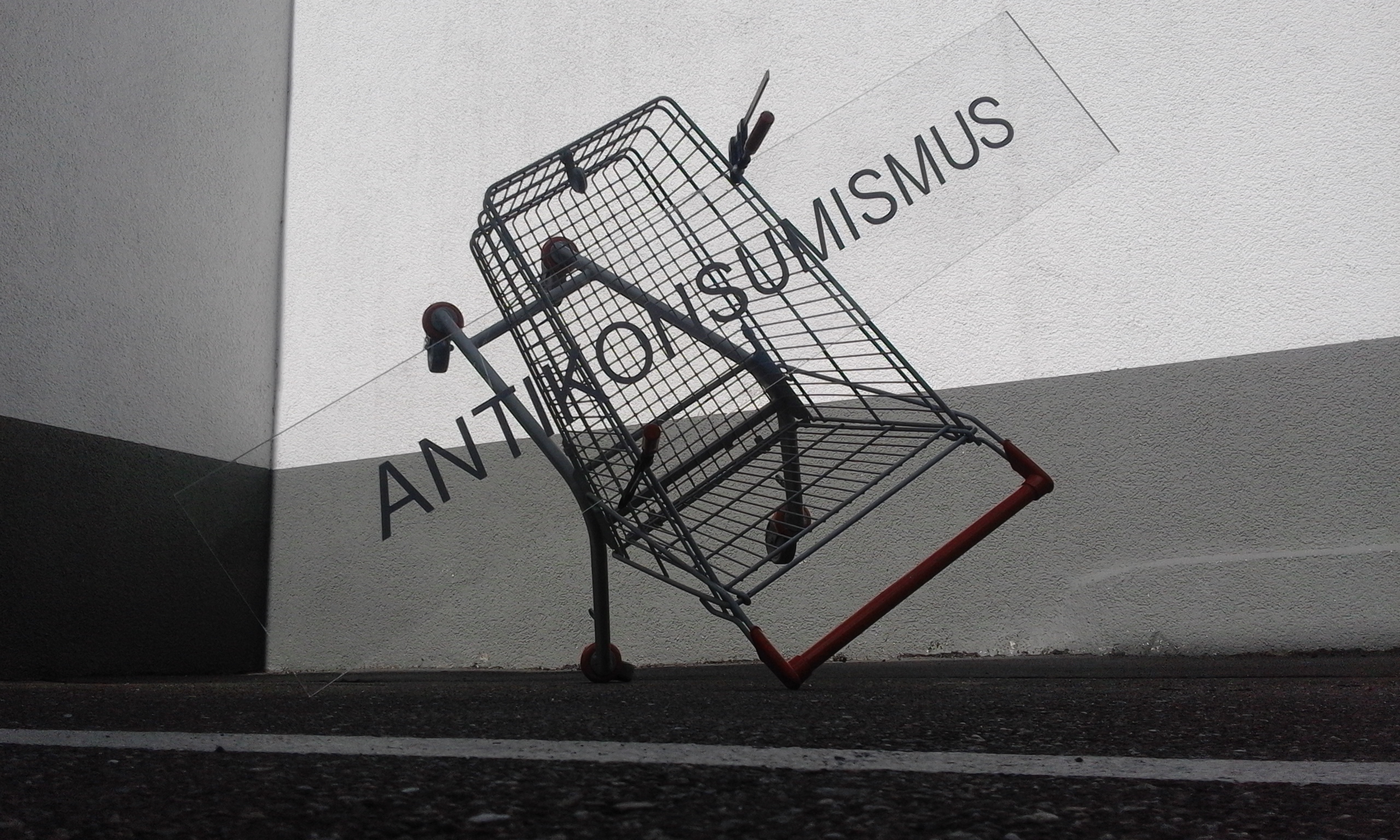Schräg aufgestellter Einkaufswagen neben der Aufschrift „ANTIKONSUMISMUS“.