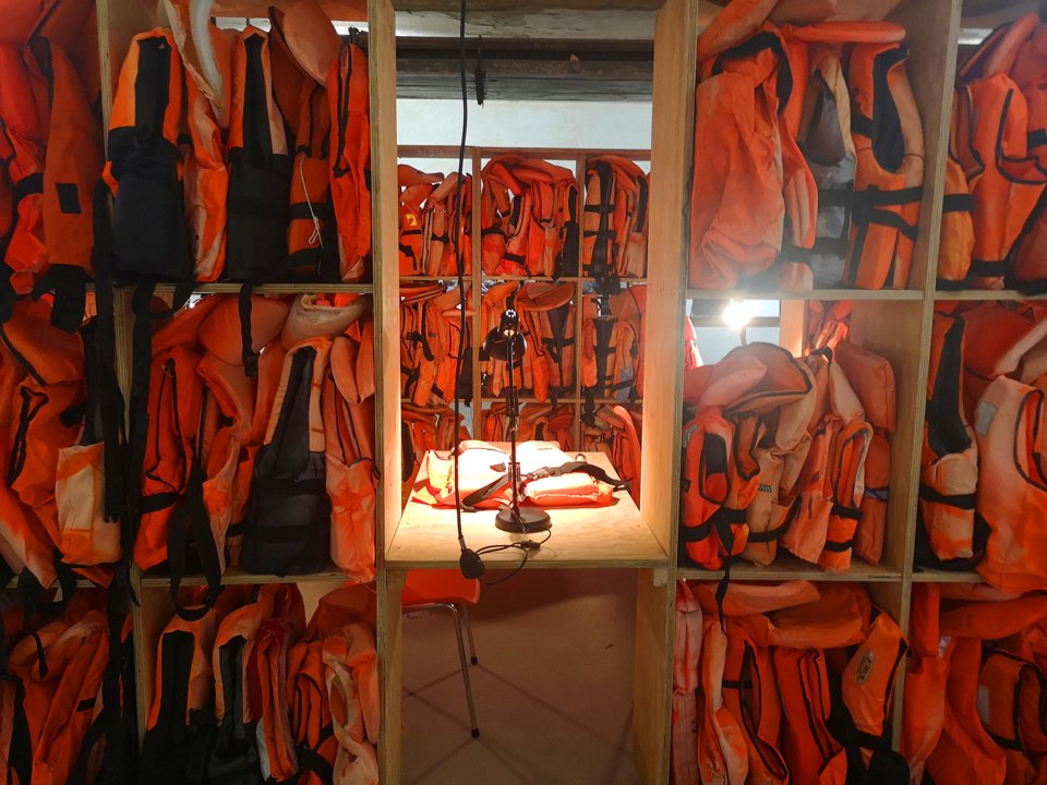 Regalfächer mit orangefarbenen Schwimmwesten, daneben ein beleuchteter Tisch und rote Stühle.