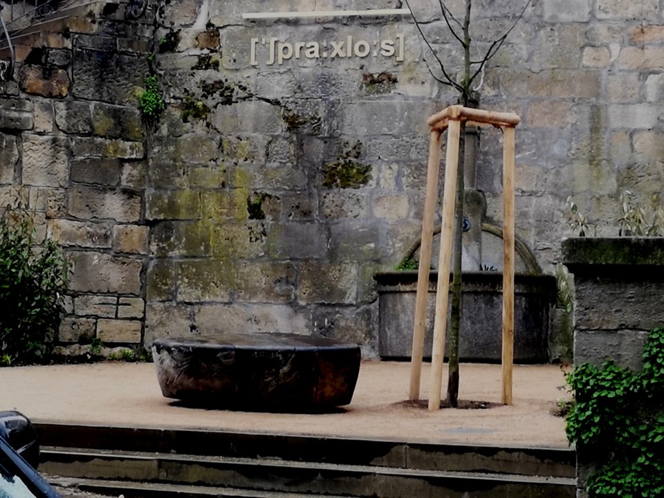 Baumstamm in einem Becken vor einer Steinmauer mit Lautschrift-Aufschrift; daneben steht ein weiteres Becken.