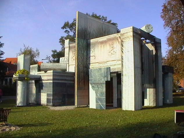 Gebäudeartige Installation aus bedruckten Architekturflächen und Säulenformen auf einer Wiese.