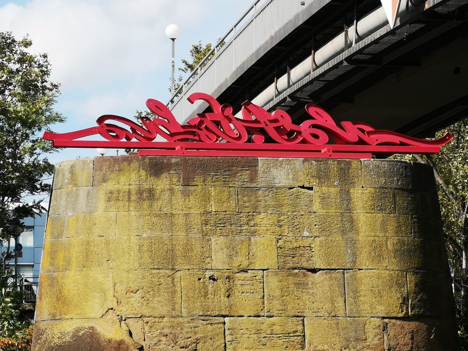 Der rote geschwungene Schriftzug liegt auf einem steinernen Brückenpfeiler über einem Fluss.