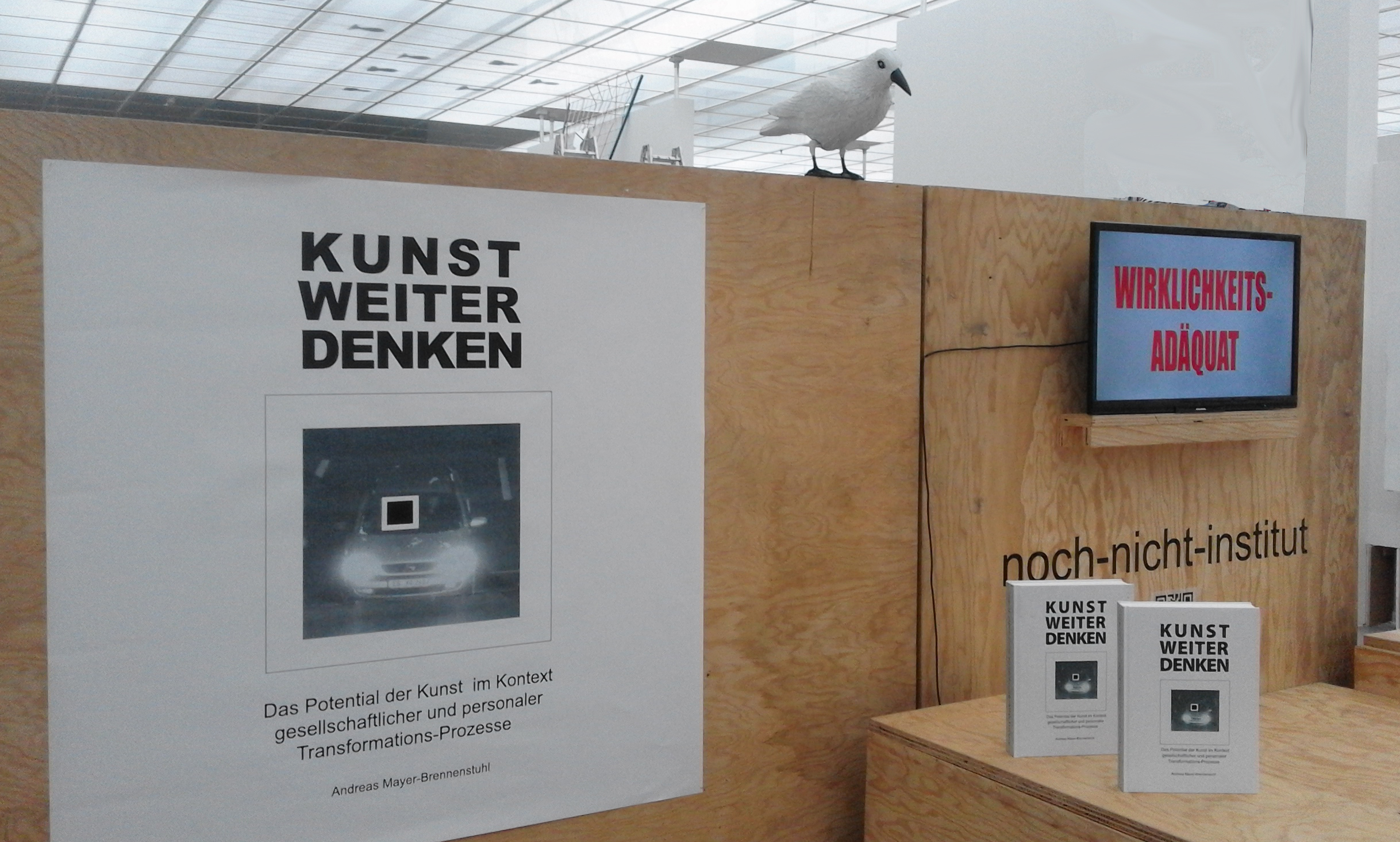 Holzwand mit dem Plakat „KUNST WEITER DENKEN“, einem kleinen Bildschirm und einem Tisch.