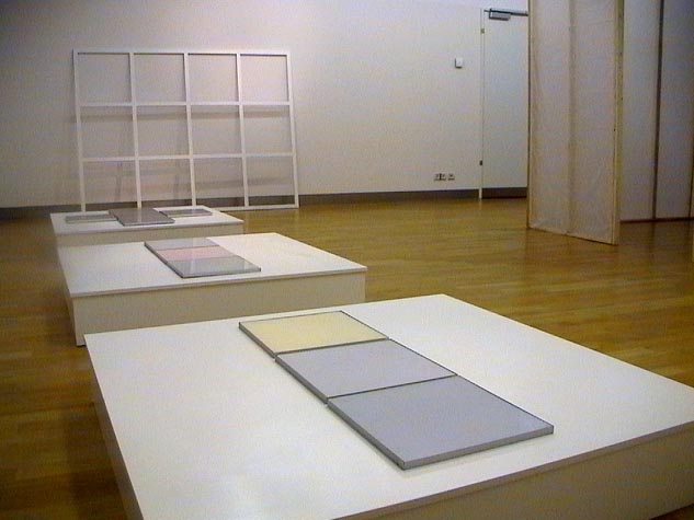 Helle Podeste mit grauen Tafeln und einem weißen Rasterrahmen in einem Ausstellungsraum.