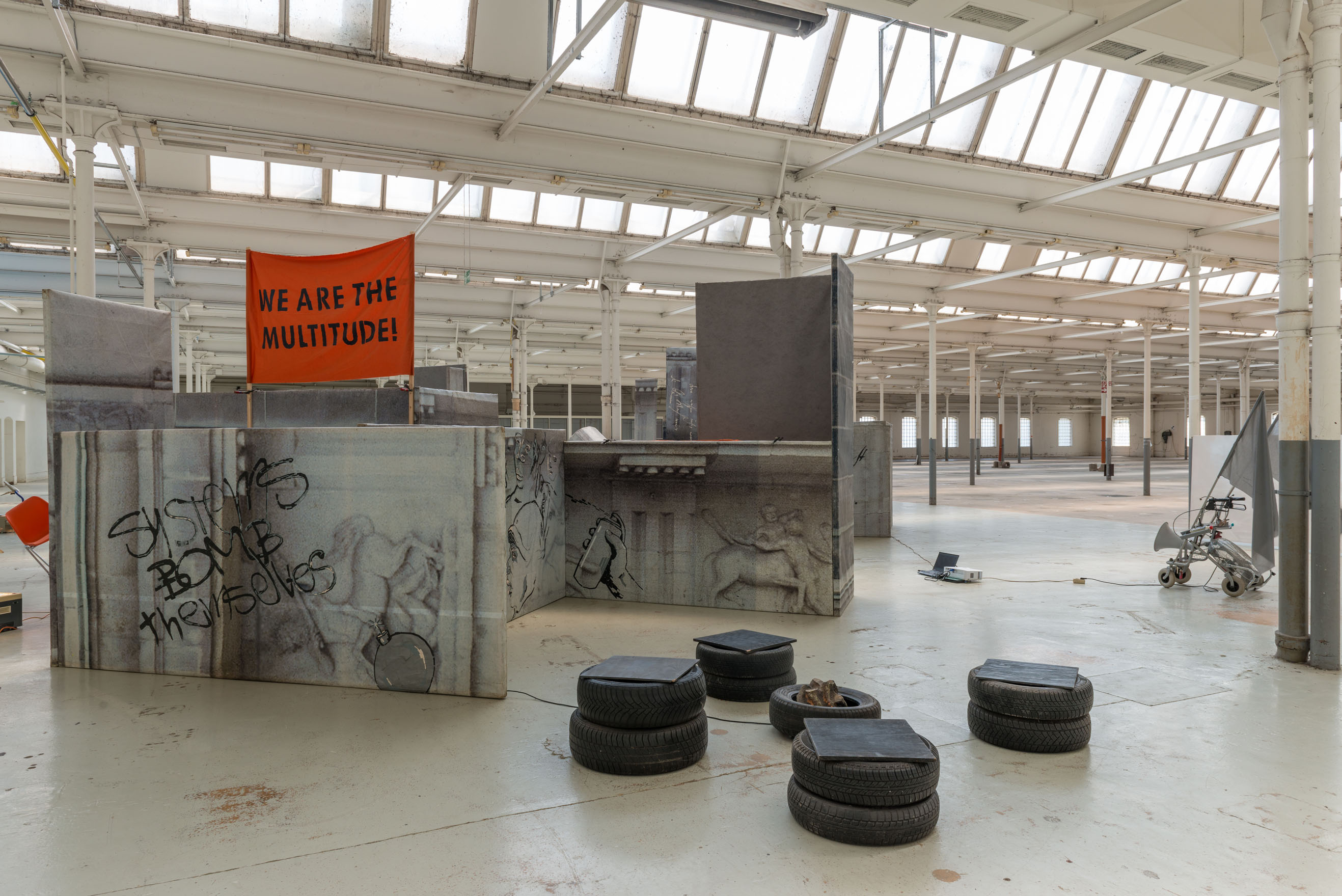 Installation aus bedruckten Architekturflächen und Reifen in einer hellen Halle; oben hängt „WE ARE THE MULTITUDE!“.