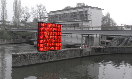 Regal mit orangefarbenen Schwimmwesten auf einer Mole vor einer Brücke und einem Gebäude.