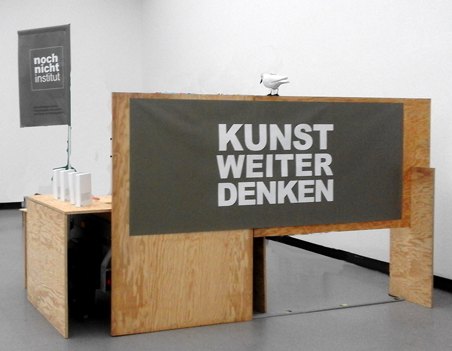 Rückansicht der Holzkonstruktion mit dem Schild „KUNST WEITER DENKEN“.