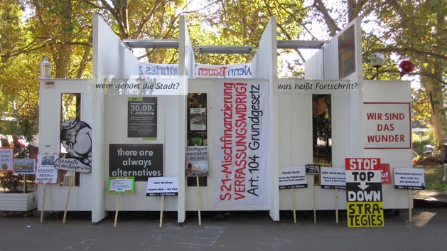 Frontansicht des weißen Pavillons mit zahlreichen Plakaten und der Frage „Wem gehört die Stadt?“.