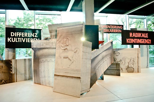 Bedruckte Architekturflächen mit Figurenreliefs und Texttafeln bilden einen Aufbau in einem hellen Raum.