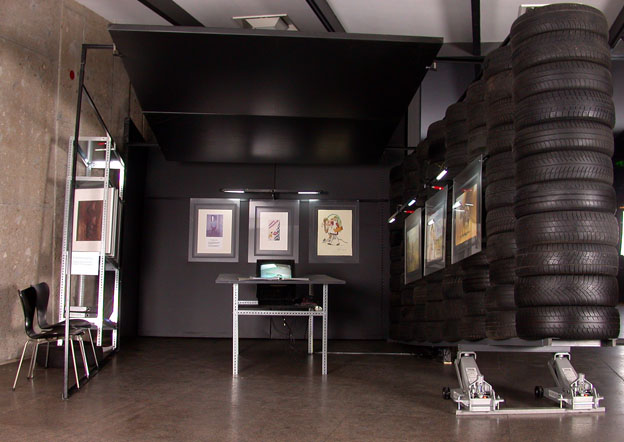 Ausstellungsraum mit hohen Wänden aus gestapelten Reifen, gerahmten Bildern und einem Tisch.