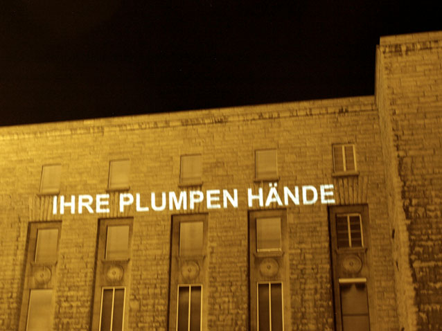 Die Worte „IHRE PLUMPEN HÄNDE“ werden nachts auf eine Gebäudefassade projiziert.