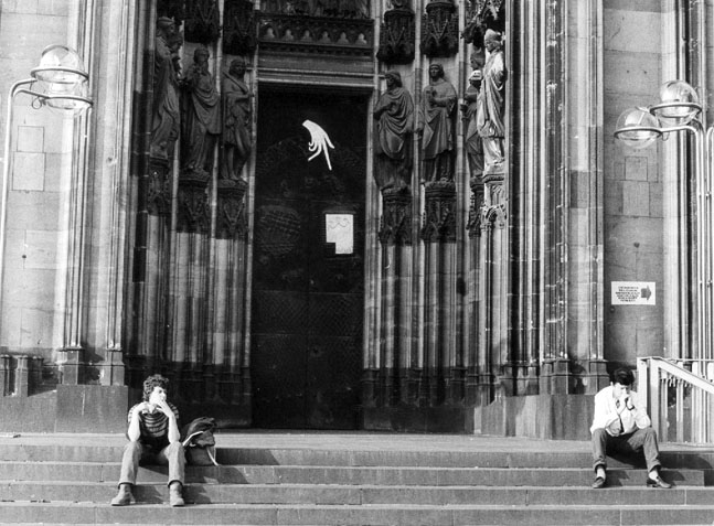 Schwarz-weiße Aufnahme eines großen Kircheneingangs mit Säulen; auf den Stufen sitzen zwei Personen.